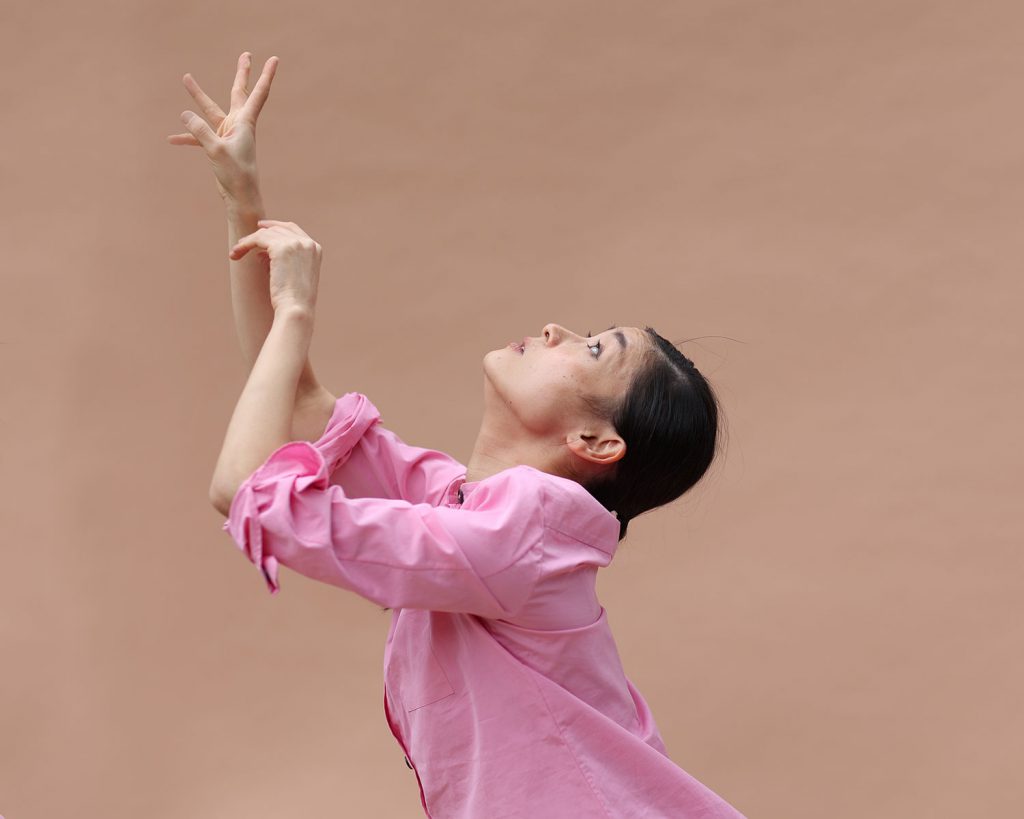写真：ピンクのシャツを着て、植物のように天に向かう手を仰ぐ北川さん