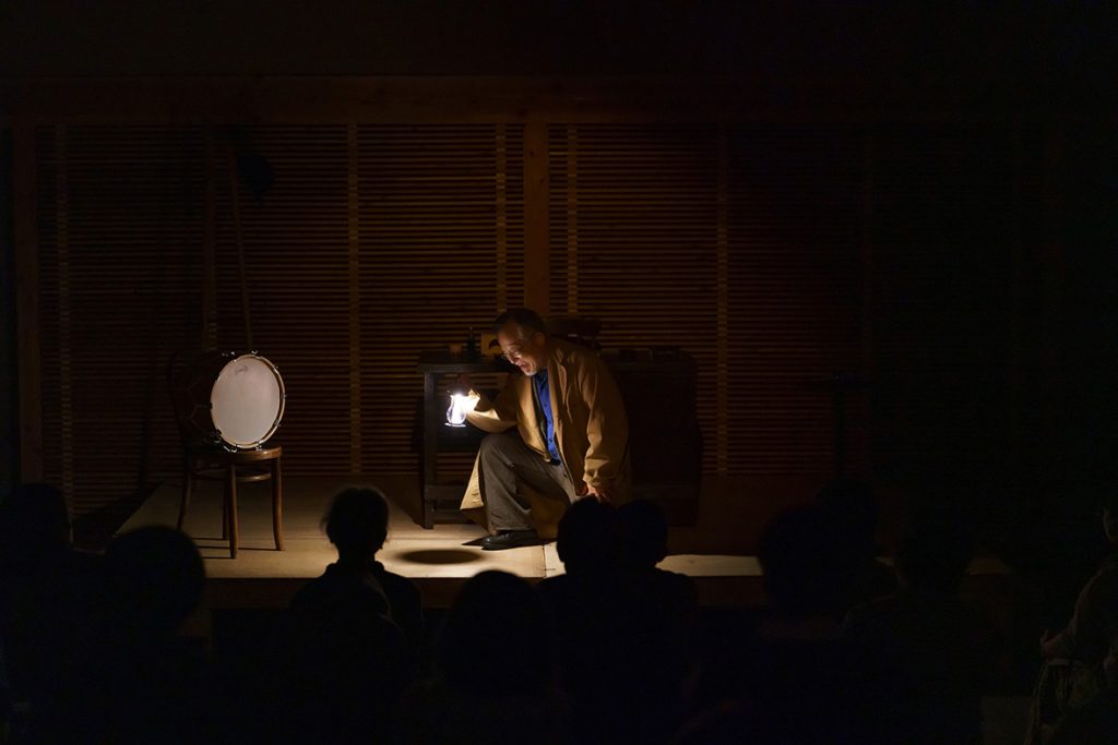 写真：暗いステージで串田さんがランプを持ってしゃがんで話している