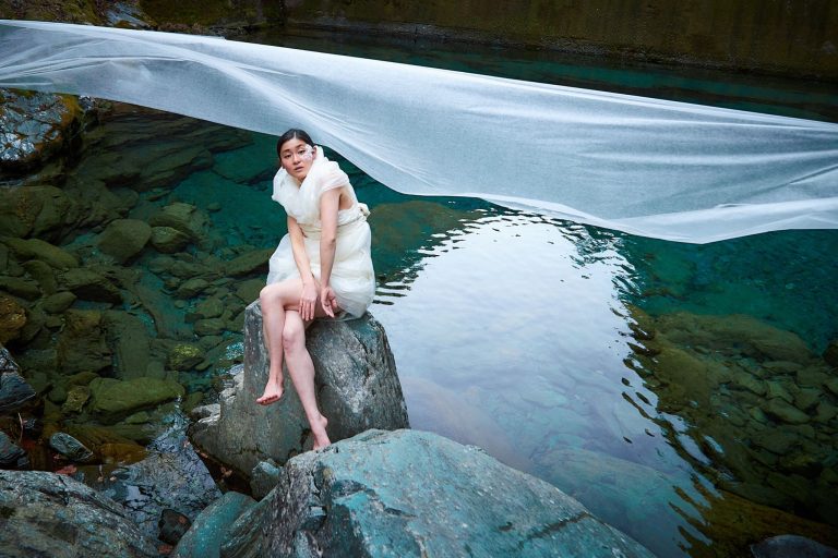 写真：川の中の岩に和紙をまとった女性が座っている。背後に薄い和紙が川の上を風のように横切っている。