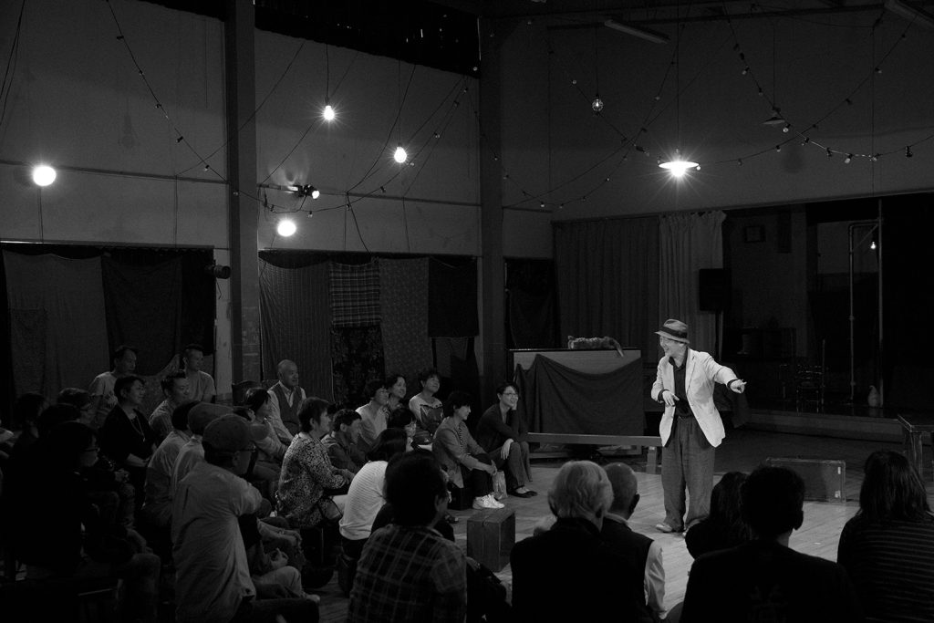 モノクロ写真：串田さんが観客に囲まれて1人で芝居をしている