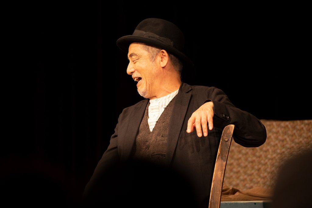 写真：串田さんが黒いジャケットと帽子を被り、椅子に座って笑っている