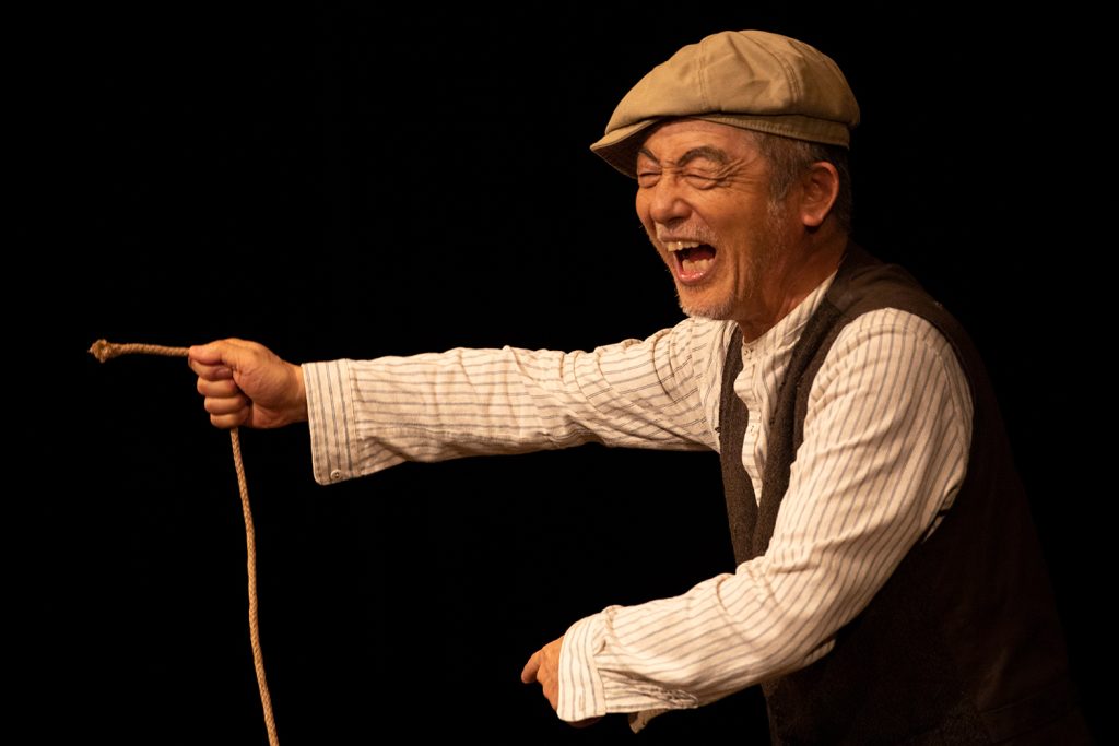 写真：ハンチング帽にベスト姿の串田さんが、ロープの端を持って口を開けている。