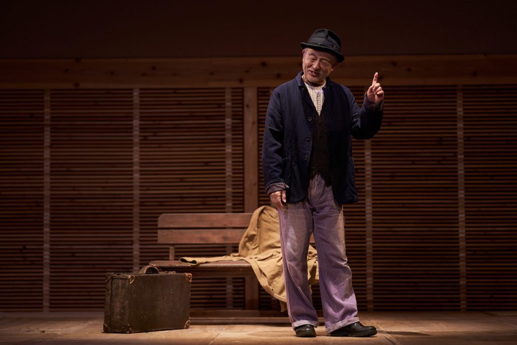 写真：1人芝居の様子。串田さんがベンチの前に立って右手の人差し指を立てている。傍らには古びたトランク。