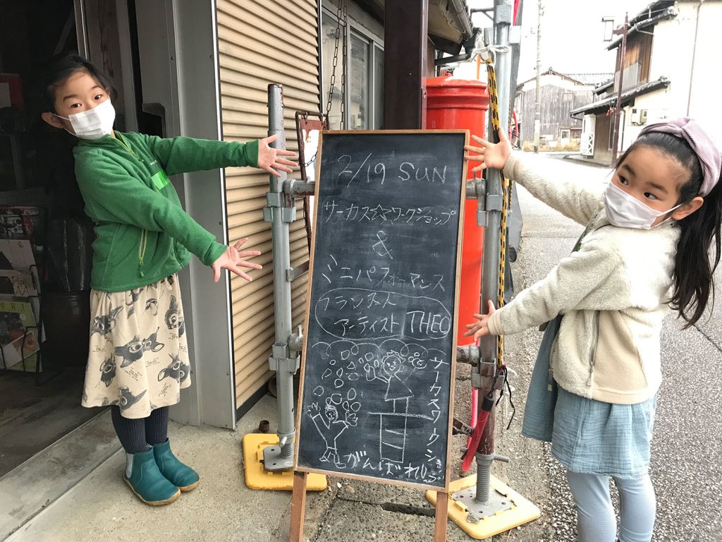 写真：2人の女の子が、イベントタイトルの描かれた立て看板を紹介している。看板は黒板でできていて、球投げをする人のイラストと、「フランス人アーティストTHEO」の文字がチョークで書いてある。