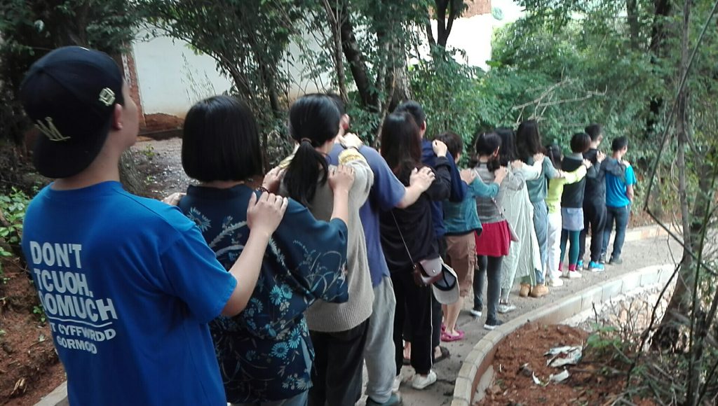 写真：一列になった人たちが、前の人に肩に手を置いて歩いている