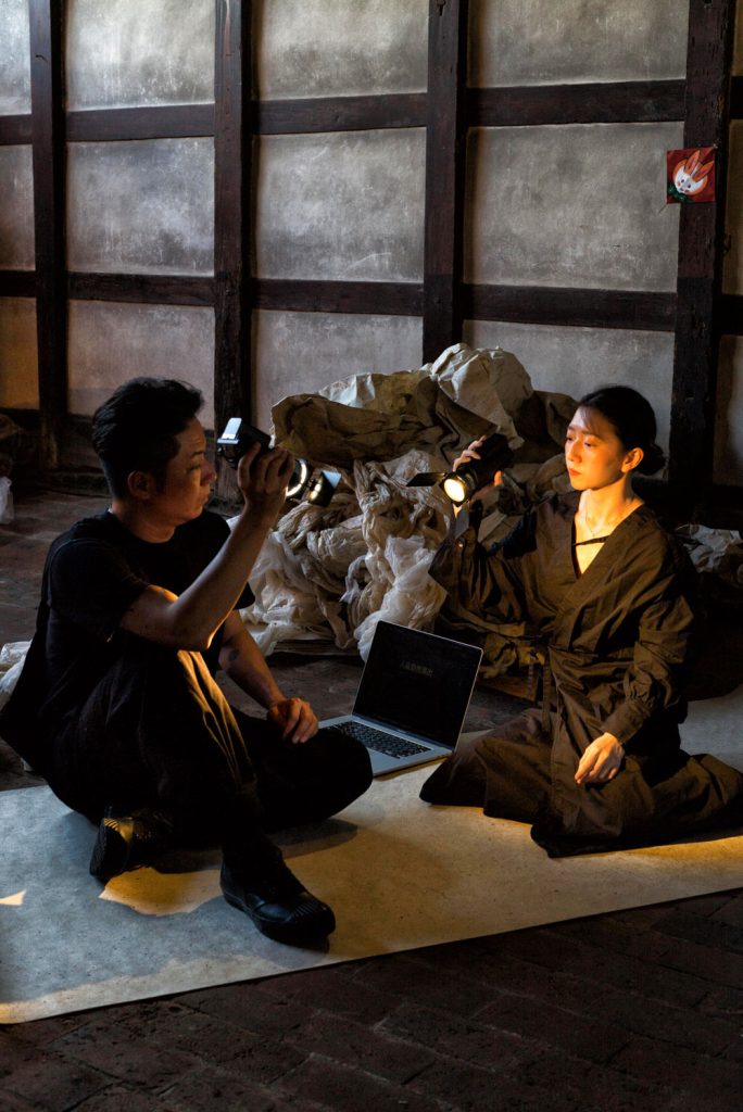 写真：男女のパフォーマーが和紙の上に座ってお互いをライトで照らしている。背後には和紙の山。