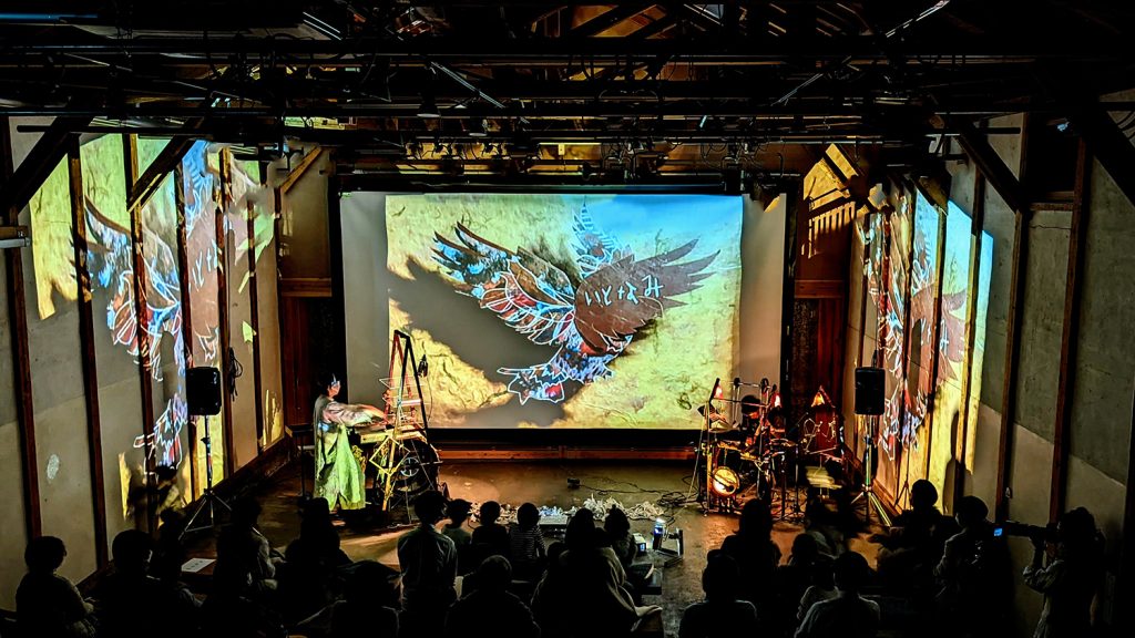 写真：舞台左右で不思議な楽器を演奏するアーティスト。中央のスクリーンと左右の壁に「いとなみ」と書かれた鳥の絵が映し出されている。