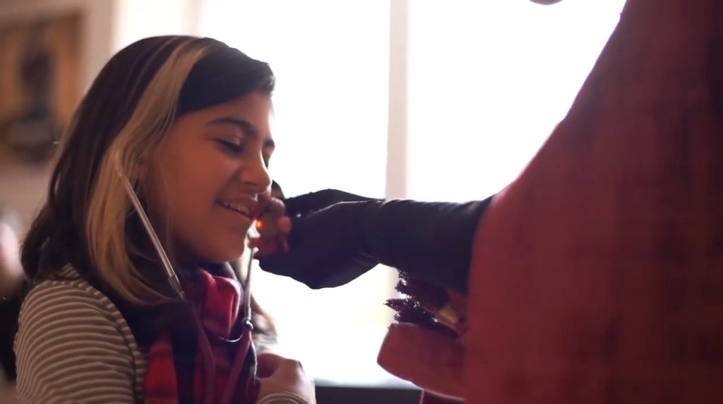 写真：聴診器をつけた女の子に、人形を近づけているアーティスト。